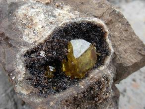 Битум, Кальцит, Сера самородная. Прозрачный кристалл серы в жеоде выполненной скаленоэдрическими кристаллами кальцита. Жеода подкрашена пленками битума.